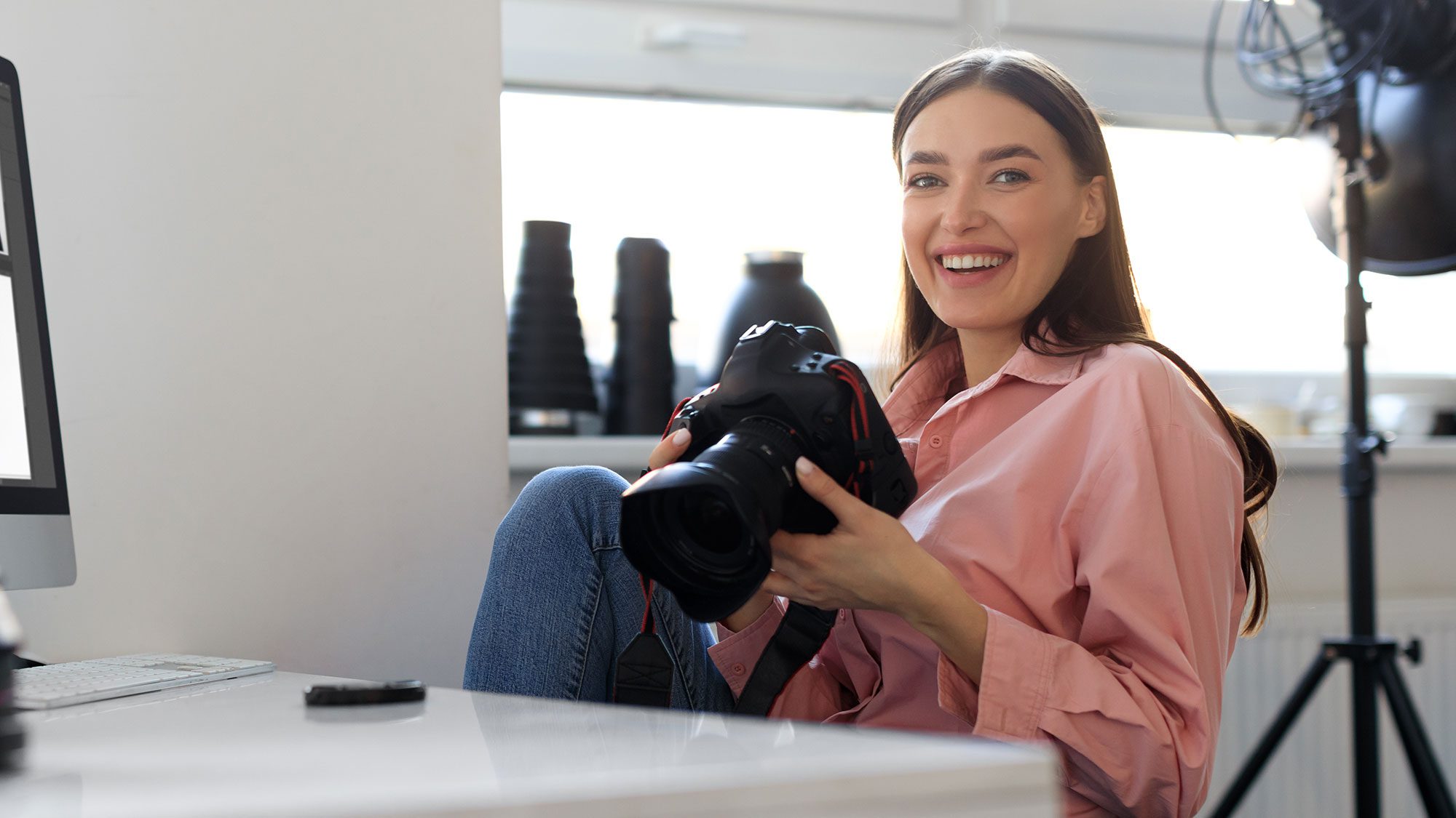 lächelnde fotografin sitzt an ihrem schreibtisch vor dem computer mit ihrer kamera in der hand